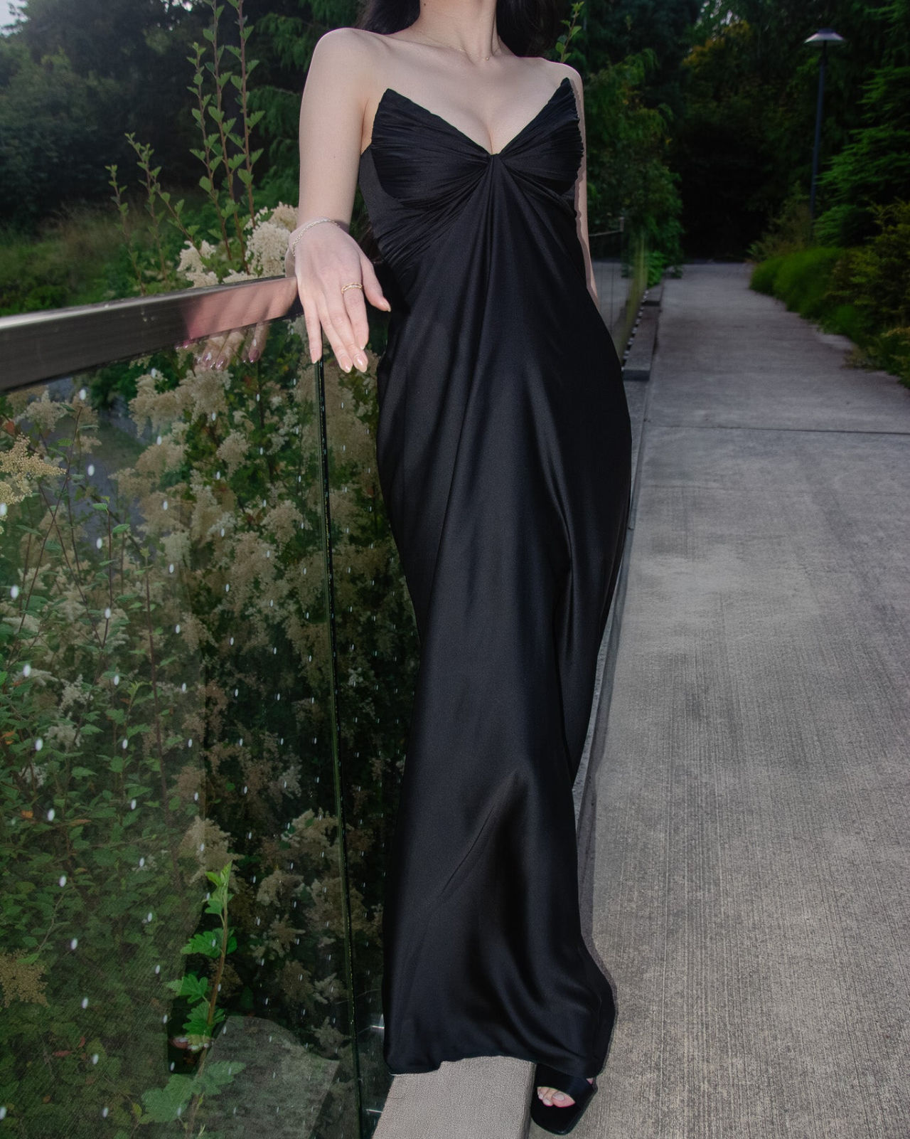 Person wearing a black dress standing on a pathway with greenery in the background